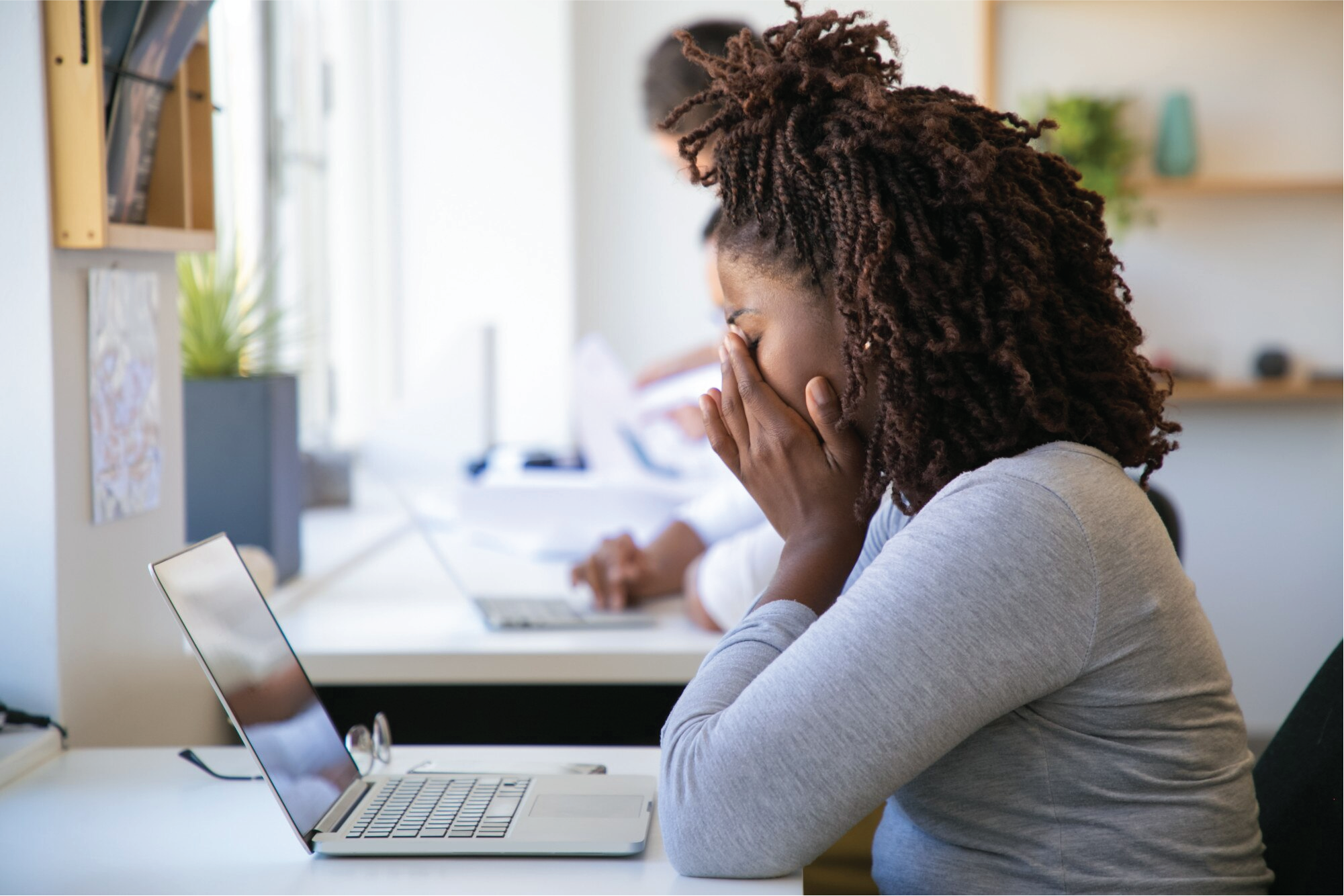 Young adult presenting female sitting at workspace with laptop open, glasses off, covering their face as if they are overwhelmed